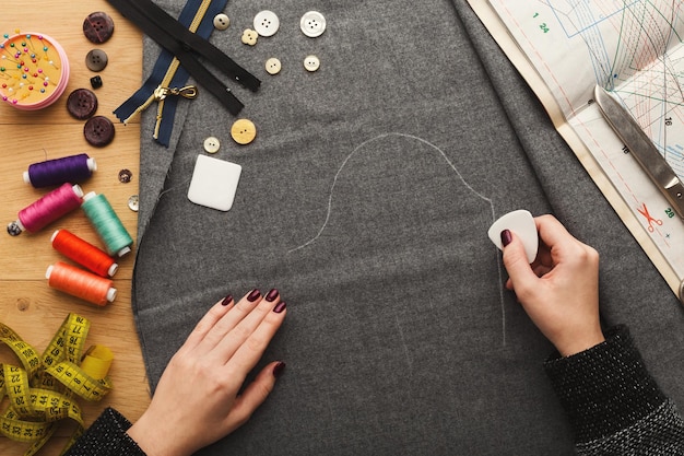 Sewing workshop or fashion designer at work. Top view on female hands marking cloth square with soap piece. Messy table with dressmaker accessories and patterns, copy space