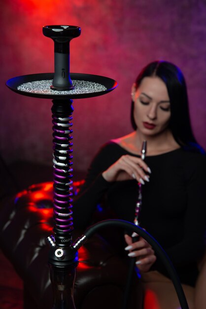 Sexy brunette girl in seductive black clothes smokes a hookah in the club