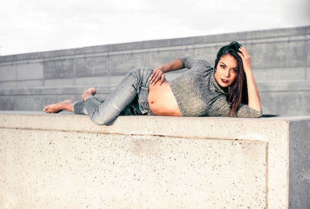 Sexy brunette in jeans and a gray sweater posing in a concrete location