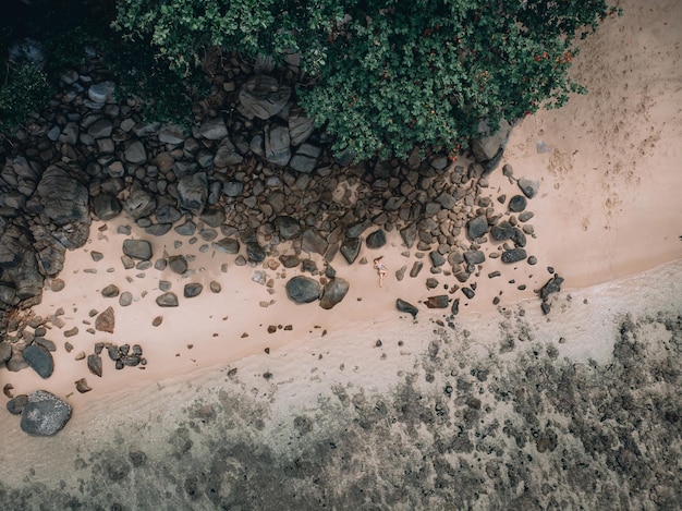 Sexy caucasian woman on the beach on the sand near the waves and stones. Beautiful woman covers her naked breasts with her hands. Phuket. Thailand. Aerial top view.