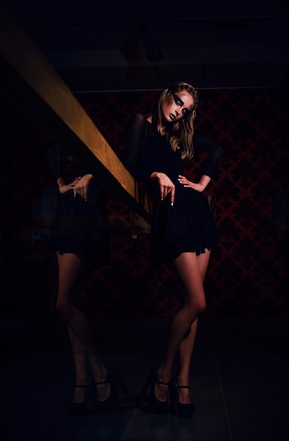 Sexy girl in black dress with gold makeup posing indoors in a dark atmosphere. reflection of feet.