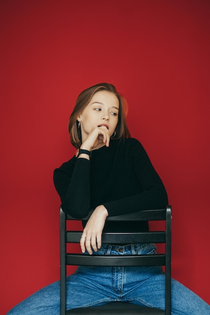 Sexy girl in casual clothes sits on a chair against a red wall wears a black sweater