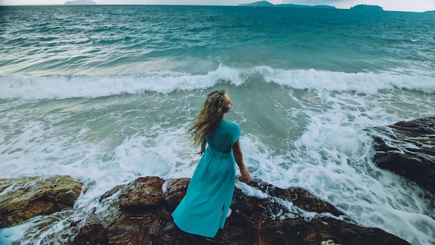 Sexy hot woman meditates relaxes on a rock reef hill in stormy morning rain cloudy sea Girl in blue swimsuit dress tunic Concept feminine relax sexual health Dark dramatic silhouette view