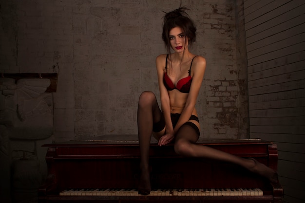 sexy leggy girl in black stockings and red bra sits on a piano in a dark room