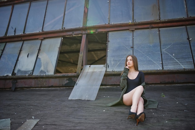 Sexy model girl wit long legs at black lingerie outfit body swimsuit combidress and jacket posed at the roof of abadoned industrial place with windows