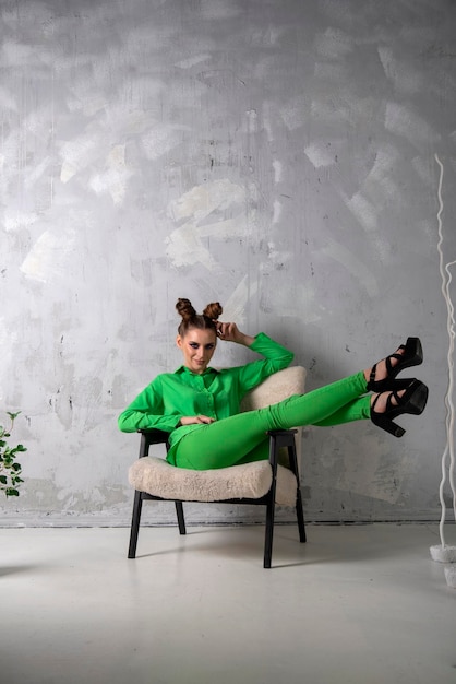 Sexy slender girl in stylish green suit and on heels sits on chair on gray wall background in studio Vertical frame