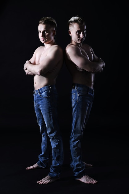 Sexy strong twin brothers with a naked torso in jeans, stand back to back. hard shadow