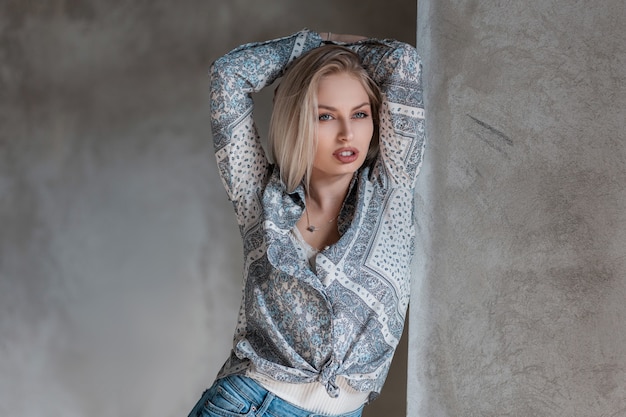 Sexy stylish young blond woman in a fashionable shirt with a pattern in a T-shirt in jeans posing in a studio near a vintage gray concrete wall