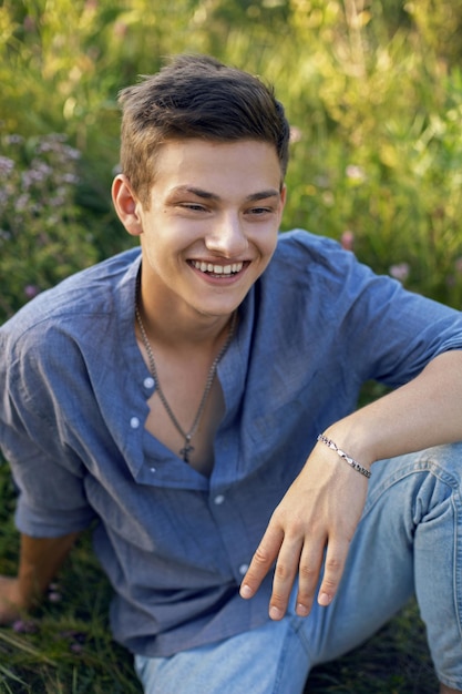 sexy teen guy sitting on the grass in nature in a shirt and jeans barefoot and looking at the camera       barefoot