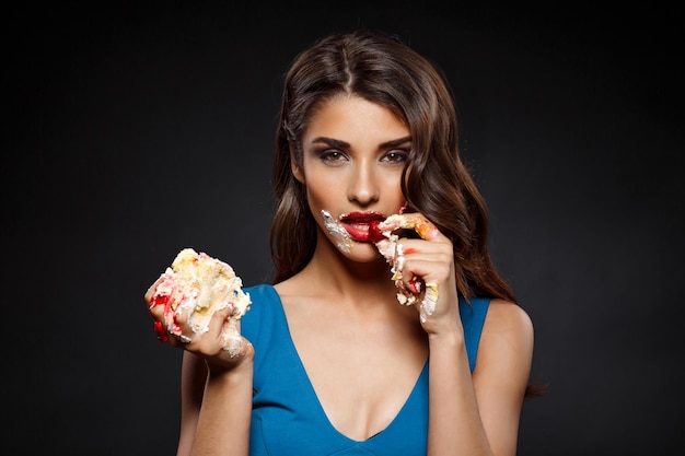 Sexy woman in blue dress eating  piece of cake over black wall