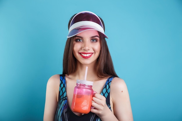 Sexy woman in swimsuit and plastic visor holding glass of fresh summer drink on blue background