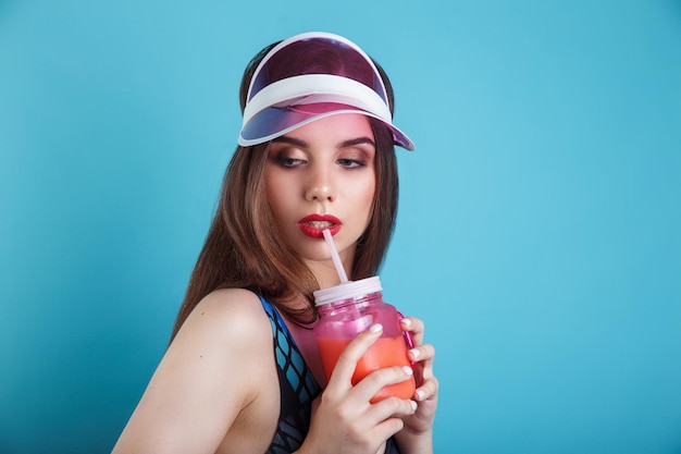 Sexy woman in swimsuit and plastic visor holding glass of fresh summer drink on blue background