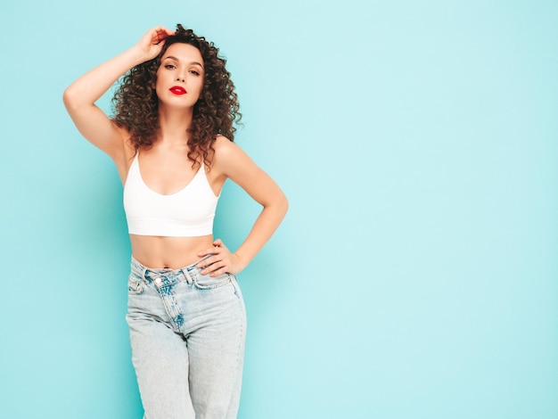 Sexy young beautiful smiling female in trendy summer hipster clothes carefree woman posing near blue wall in studio Positive brunette model with afro curls hairstyle Cheerful and happy