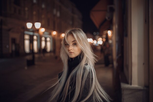 Sexy young beauty woman posing over night city illuminated street background. Vacation holidays. Young stylish woman blonde with long hair