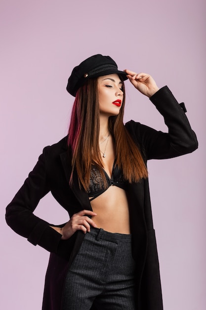 Sexy young woman in long fashionable jacket in stylish underwear in trendy pants with puffy red lips straightens stylish black cap in the studio near the wall. Modern girl fashion model indoors.