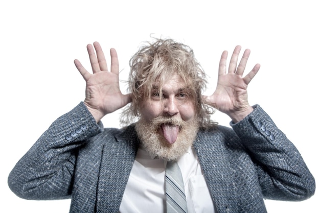 Shaggy adult grayhaired man with a beard wearing a gray suit shows tongue and holds hands near ears Isolated