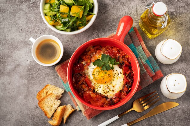 Shakshuka in a red pan, coffee, bread  and salad in a bowl, gray background, copy space