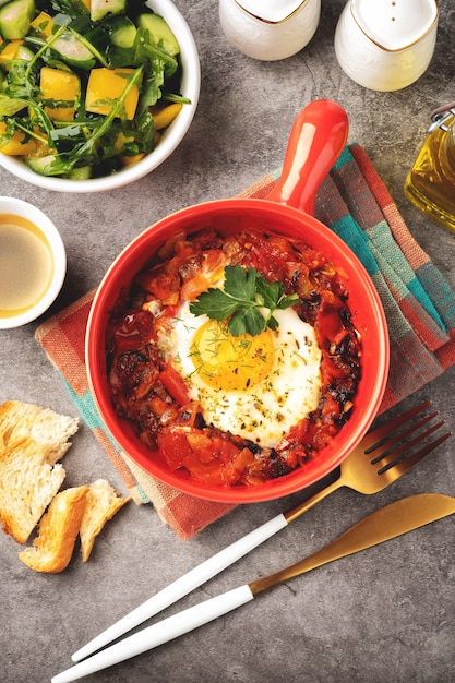 Shakshuka in a red pan on a gray background, breakfast concept with fried egg, salad and coffee