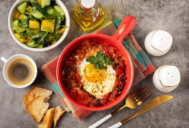 Shakshuka in a red pan on a gray background, breakfast concept with fried egg, salad and coffee