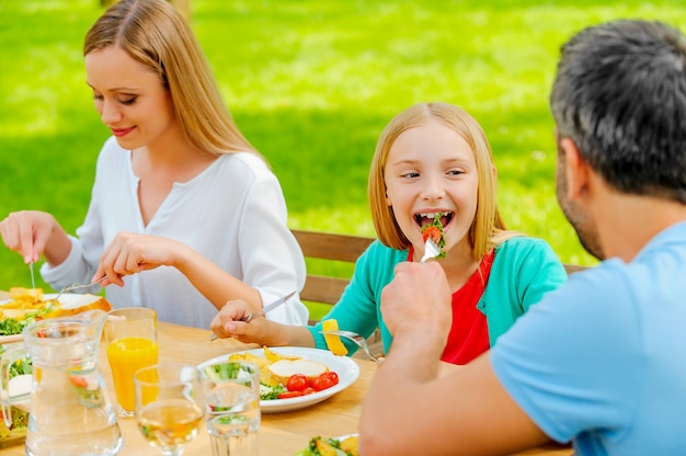 Sharing food with the nearest. Young man feeding his daughter with salad while sitting together at the dining table outdoors