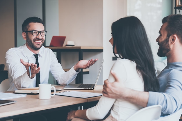 Sharing good news. Happy young couple bonding to each other and listening to cheerful mature man sitting at the desk in front of them and gesturing