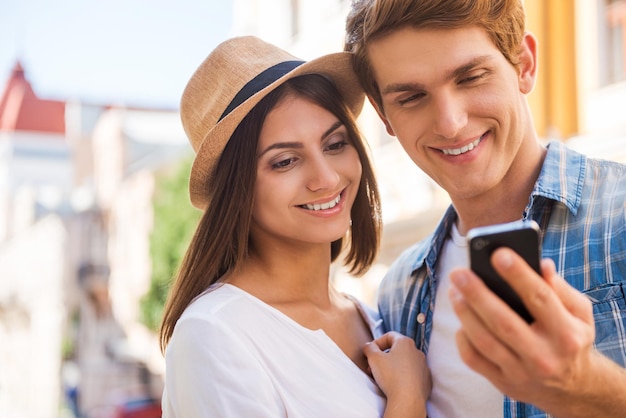 Sharing their pictures with friends. Beautiful young loving couple standing close to each other while man holding mobile phone and smiling