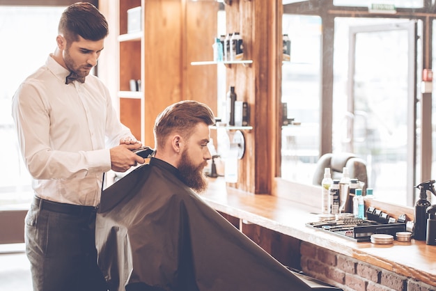 Sharp edges. Side view of young bearded man getting haircut by hairdresser with electric razor while sitting in chair at barbershop