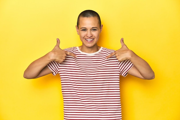 Shaved head woman in red striped tee yellow backdrop surprised pointing with finger smiling broadly