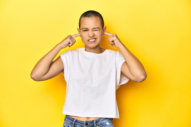 Shaved head woman in white tank top yellow backdrop covering ears with fingers stressed