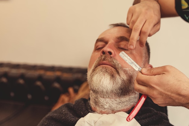 Shaving with a straight razor in a barbershop A bearded old man being shaved in a barbershop