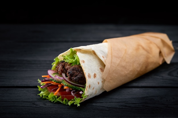 Shawarma rolled in lavash, moist grilled meat with onion, herbs and vegetables on wooden black background.