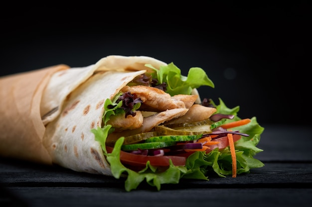 Shawarma rolled in lavash, moist grilled meat with onion, herbs and vegetables on wooden black background.