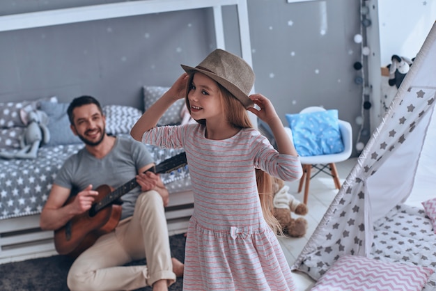 She is his little princess. Young father playing guitar for his little daughter and smiling while spending free time at home