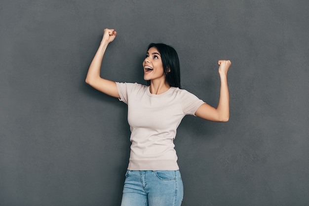 She is a winner! Happy young woman in casual wear keeping arms raised and looking away while standing against grey background