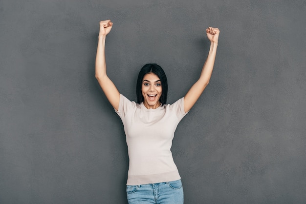 She is a winner! Happy young woman in casual wear keeping arms raised and looking at camera while standing against grey background