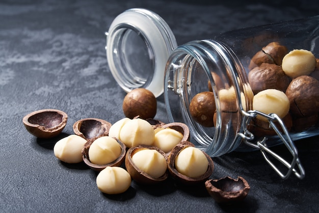 Photo shelled and unshelled macadamia nuts in an open glass jar on black table, close-up.