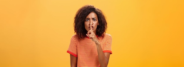 Photo shh do not disturb portrait of seriouslooking bossy and displeased cute african american girl with afro hairstyle frowning shushing at camera with index finger over mouth forbidding speak loud