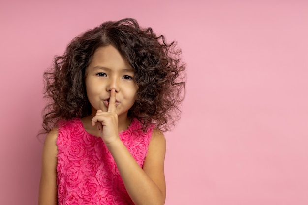 Shh. Keep our little secret. Portrait of girl in beautiful dress, with curly hairstyle, brown skin, making hush gesture, keeping index finger over mouth, standing. Copy space.