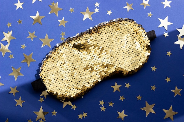 Shiny sleep mask for eyes decorated with sequins and gold stars confetti on a blue background Top view Flat lay Copy space Concept eye protection from light for good sleep and melatonin production