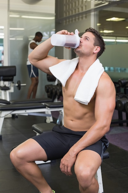 Shirtless bodybuilder drinking protein drink sitting on bench