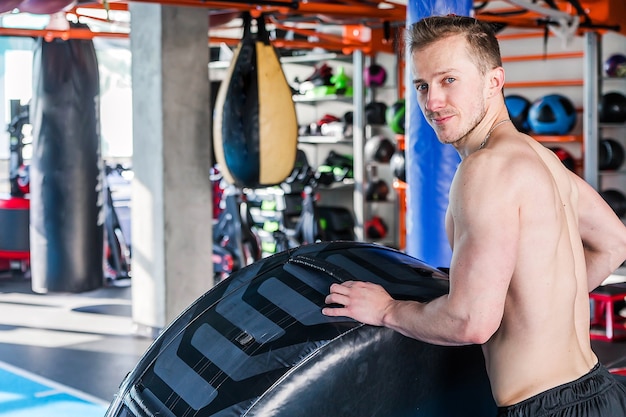 Shirtless man flipping heavy tire at gym. View from his back. Sportsman look at camera