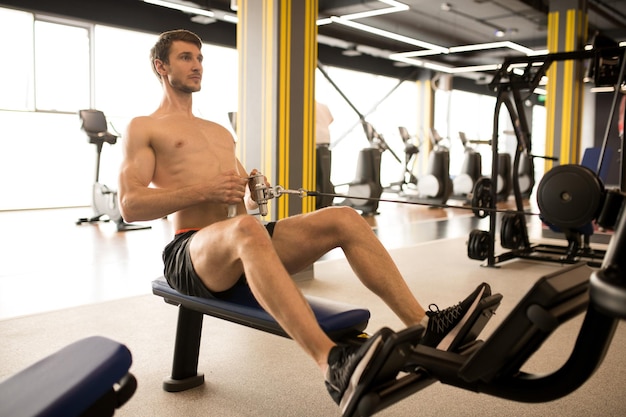 Shirtless muscular athlete sitting and pulling the weight on himself while training on rowing machin