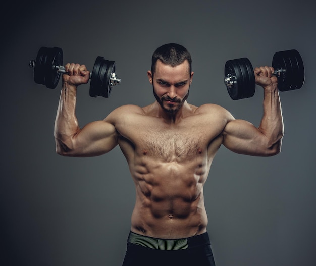 The shirtless muscular male holds dumbbells with arms up on grey background.