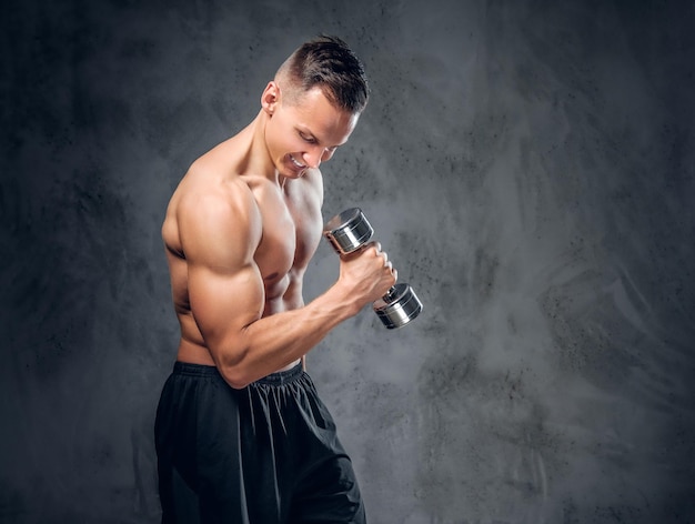 The shirtless muscular male holds a set of dumbbells over dark grey vignette background.