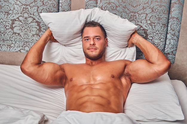 Shirtless sexy muscular male model lying alone on his bed in his bedroom, looking away.