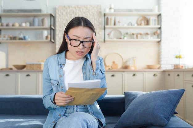 Shocked and alarmed young beautiful asian woman received divorce papers sitting at home on the couch