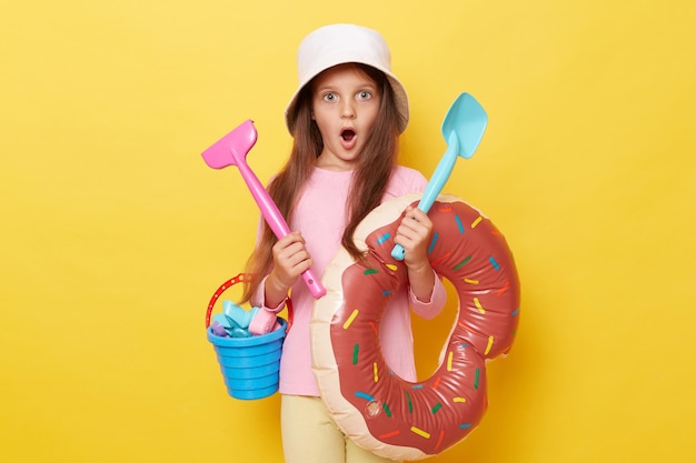 Shocked amazed little girl in panama holding sandbox toys bucket and rubber ring isolated over yellow background posing with scoop and rake keeps mouth widely opened