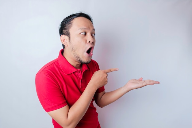 Shocked Asian man wearing a red tshirt pointing at the copy space beside him isolated by a white background