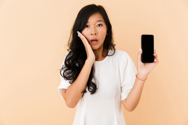 Shocked asian woman posing isolated over beige wall space showing display of phone.