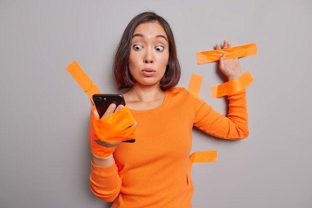 Shocked Asian woman stares at modern smartphone wrapped in plaster adhesive band being in captivity wears orange jumper stuck to grey studio wall